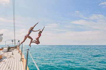 Carefree excited young women jumping from boat to swim in the ocean. Two friends on a holiday cruise together jumping from boat into the ocean to swim. Excited young women jumping off boat together - Powered by Adobe