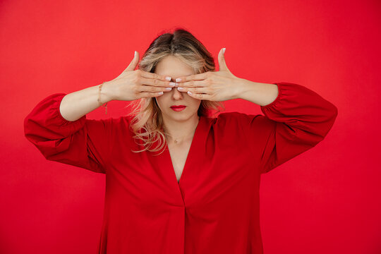 Portrait of hiding woman in red outfit with perfect bright makeup closing eyes by hands isolated on red background. Avoiding problems, fear, giving under stress. Fashion and beauty industry