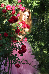 Art portrait of a Girl on a background of roses. Girl and rose bushes. Beautiful young Woman with long hair posing near roses in the garden. Rose Garden. Portrait of young Woman in the spring time. 