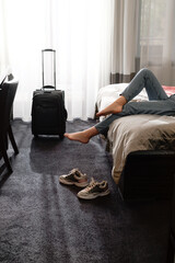 woman taking off footwear in a hotel room on the bed. Tourist relaxing on hotel room after travelling with suitcase. Female having rest after long trip with language Dark silhouette  - obrazy, fototapety, plakaty