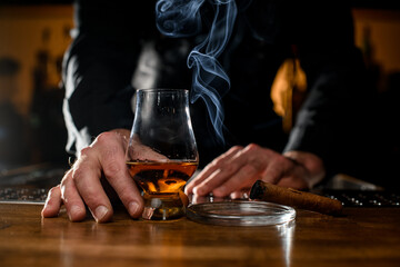 great view on male hand holding glass with cognac and ashtray with a smoking cuban cigar nearby