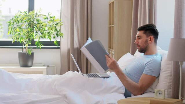 people, technology and remote job concept - man with laptop computer and folder working in bed at home bedroom