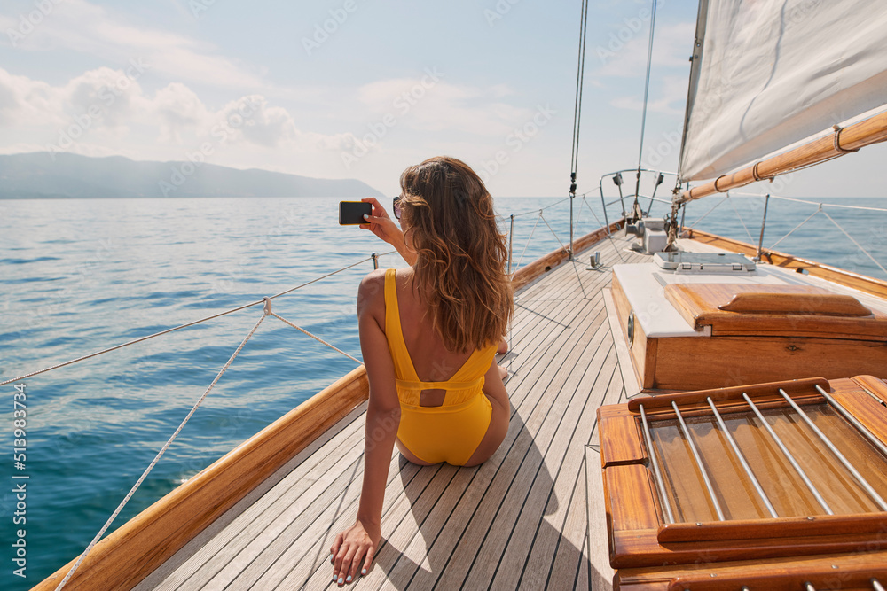 Wall mural young woman on holiday cruise in yellow swimwear taking photos of the ocean on her smartphone. woman