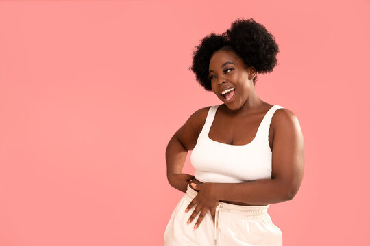 Attractive female model with dark skin and afro hairstyle posing on pink pastel studio background, smiling and showing her belly. Diet concept. Body positive, conscious.