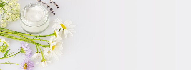 Concept of wild-harvested beauty and natural cosmetics based on a wild plant. Glass jar with organic creams and wild flowers on a white background with copy space. Soft focus style, banner size