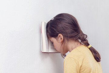 Sad and tired caucasian girl with dyslexia holds a book with her forehead. The child learns to speak and read correctly - obrazy, fototapety, plakaty