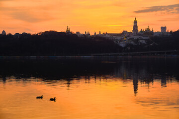 Fototapeta premium Kiev-Pechersk Lavra and city lights. Silhouette panorama at sunset of the colorful sky over the Dnieper river with passing ducks in Kyiv