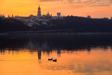 Fototapeta na wymiar Kiev-Pechersk Lavra and city lights. Silhouette panorama at sunset of the colorful sky over the Dnieper river with passing ducks in Kyiv