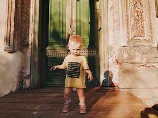 One year or 12 months old baby boy standing on a beautiful 
wooden porch of an old Russian 19th...