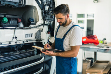 Truck mechanic writing notes in notebook during working in modern workshop. Young concentrated caucasian bearded man.Truck service, repair. Garage interior with tools and equipment