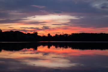 Sunset over the river. Scenic summer landscape with trees silhouettes and clouds reflections in the water