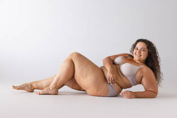 Fototapeta premium Portrait of beautiful woman with plum figure posing in underwear isolated over grey studio background. Self-acceptance, self-love