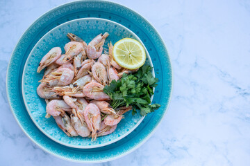 Сooked shrimp with parsley and lemon in a blue ceramic bowl on a white marble table background. Healthy Mediterranean seafood. Top view, flat lay, copy space