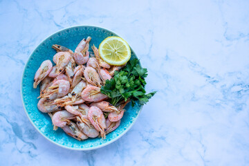 Сooked shrimp with parsley and lemon in a blue ceramic bowl on a white marble table background. Healthy Mediterranean seafood. Top view, flat lay, copy space
