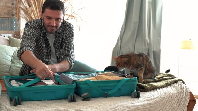 Smiling man packs winter themed cyan bag for holiday. Vacation preparation, careful bag packing. Millennial generation Y travel blogger pack suitcase and petting cat. loft design room. 4k footage