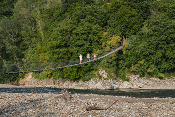 View of the suspension bridge and people passing through it through the mountain river. Wooden suspension bridge in a mountainous area on a sunny day. Suspension bridge over the river.