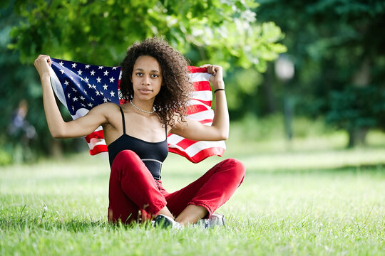 Young African american woman at nature background with an American USA flag on her shoulders. USA resident, US citizen. Immigration concept