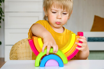 Little blond hair boy in clothes made of natural fabric plays with rainbow colored wooden toys at...