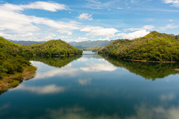 Fototapeta premium Aerial view of river among mountains and hills with a reflecting blue sky and clouds. Randenigala reservoir, Sri Lanka.