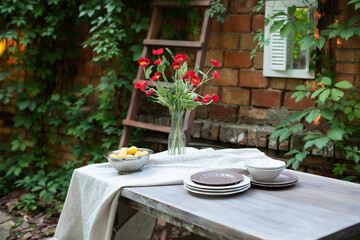 A bouquet of red poppies in a glass vase. Flowers in a vase at home. Interior Terrace, patio. Wooden table and plates for garden party or dinner. Table for lunch outside in garden in yard of house.	