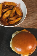 Overhead shot, tasty wedge fries in a white bowl, accompanied by a hamburger.