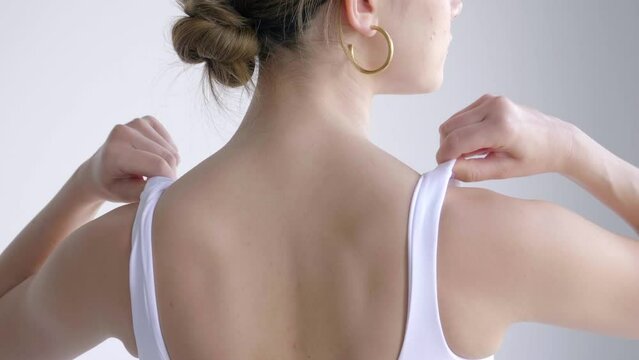 Back View Of Lovely Young Woman Facing White Wall Wearing White Tank Top Adjusting The Strap.- Medium Closeup, Slowmo