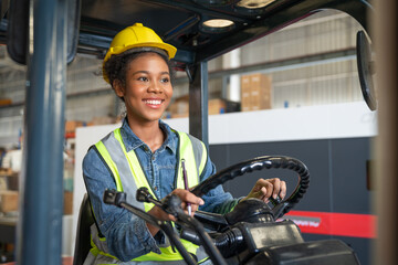 Young African American female worker forklift driver in safety vest with yellow helmet smiling and happy while working in industry factory logistic shipping warehouse.