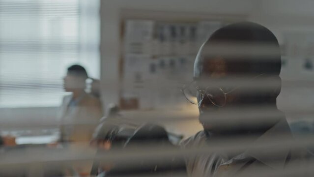 Chest-up view through window with horizontal blinds of young African American male agent taking pictures on professional camera from cover in office
