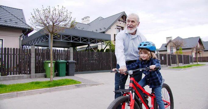 Happy old grandfather teaching small cute smiled grandson in helmet to ride a bike at street near house. Grandpa day. Having fun together. Little boy learning riding bicycle and senior man helping.