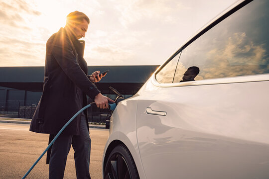 An elegant businessman charging a new white electric car, in the parking of the trade center at sunset