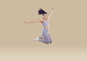 Cheerful positive woman in sportswear having fun jumping high on beige background. Full length of...