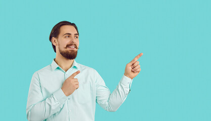 Advertise here. Happy male customer presenting new product, pointing finger at copy space on light blue background. Smiling millennial man with beard and mustache wearing shirt advertises recommends