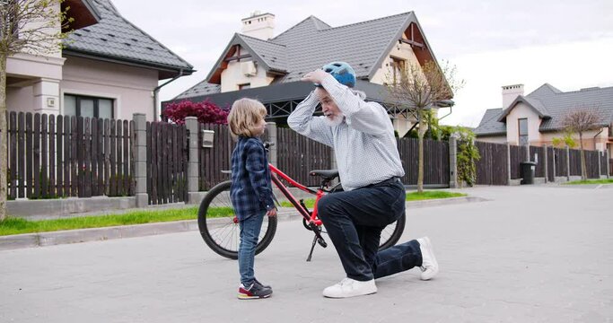 Caucasian grandfather teaching small cute grandson riding a bike. Grandpa putting on helmet on head of little boy. Outdoor. Senior man learning how to ride bicycle a kid.