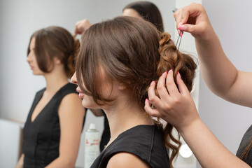 The hairdresser makes a hairstyle to a young attractive woman with a beautiful make-up in a black dress in a beauty salon.