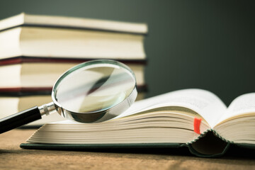 Magnifying glass on the opened book on the table, with pile of books on background - obrazy, fototapety, plakaty