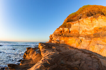 Fototapeta na wymiar Beach Ocean Coast Large Sandstone Rocky Cliff In Morning Blue Sky Sunlight Colors A Scenic Landscape.