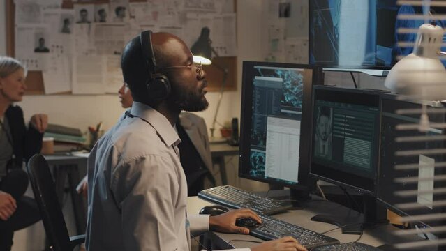 Side view of young Black man using computer, searching federal criminal database, two female detectives sitting and talking on background in office