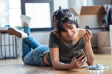Young beautiful woman with mobile phone while eating a piece of chocolate lying on the floor in her...