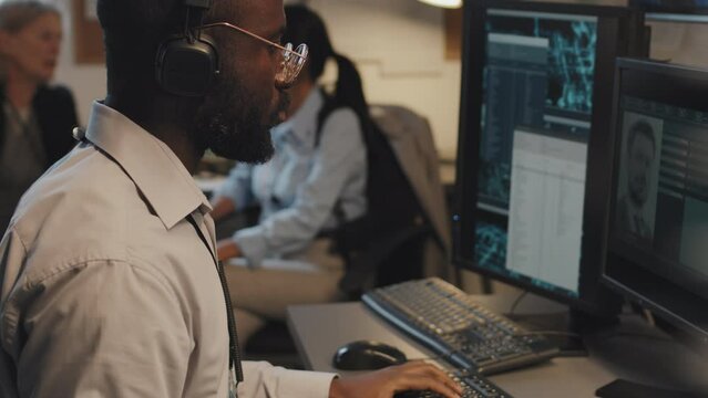 Side view tilting up of young Black man using computer, searching FBI criminal database, two blurred female detectives sitting and talking on background in office