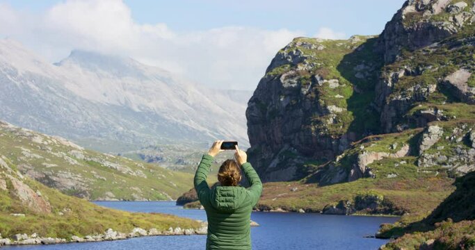 Rear view of a young woman using a smartphone to take a picture of a mountain. Unrecognizable woman taking a picture with her phone while hiking through nature on an adventure travel vacation