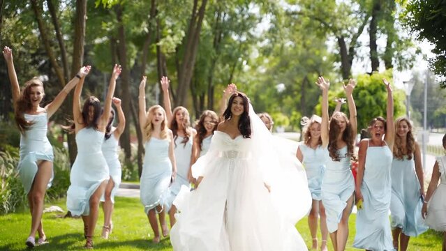 Beautiful bride, pretty bridemaids in pale blue dresses walk, run in park cheering waving hands. Woman with long airy bridal veil in elegant fluttering white dress partying with friends. Slow motion.
