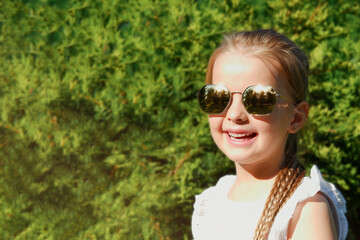 Portrait of happy pretty child girl in sunglasses outdoors. Green plant background.Copyspace.Summertime