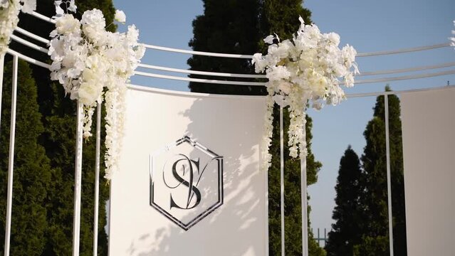 Decorated wedding ceremony arch background with initials of bride anf groom name. Wedding arch decorated with the white flowers, greenery outdoors. Newlyweds family logo of two first letters of names.