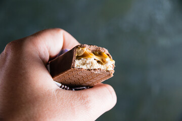 Delicious chocolate bar with caramel and peanuts close up in female hand on dark background with copy space