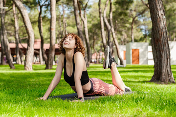 Naklejka na ściany i meble Beautiful redhead woman wearing sportive clothes standing on city park, outdoor doing yoga, stretching on yoga mat. Practicing stretching high cobra asana pose.