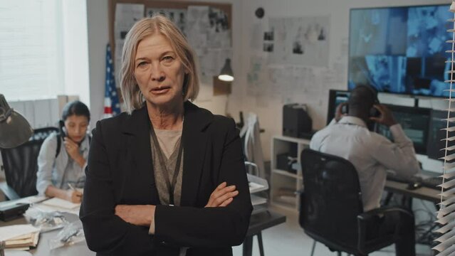 Waist-up portrait of mature Caucasian female prosecutor in formal suit crossing arms on chest, standing in office at daytime, looking on camera