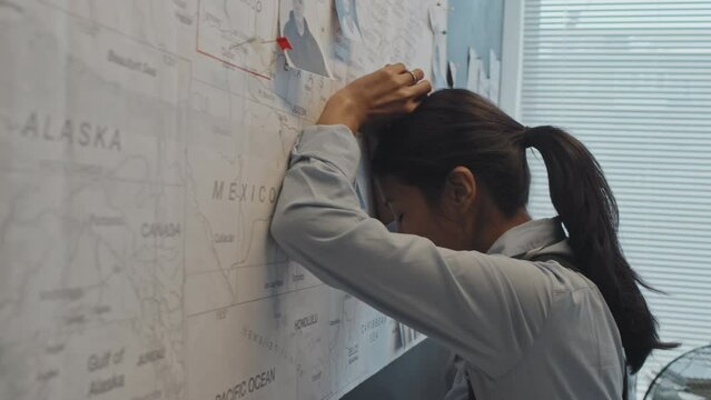 Side view chest-up of tired young Hispanic female detective looking at pinned pictures of criminals on map on wall in office at daytime