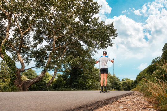 A sporty man in a helmet trains on roller skis on a country road, rear view. Biathlon race with ski poles. Low angle view. Blue sky and beautiful nature along the road. The concept of summer workout