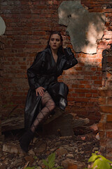 Fototapeta na wymiar a young pretty woman in a black leather coat, raincoat in a park near a destroyed building