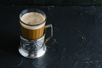 Aromatic cappuccino coffee in a glass cup with a vintage coaster on a dark concrete background with copy space.
